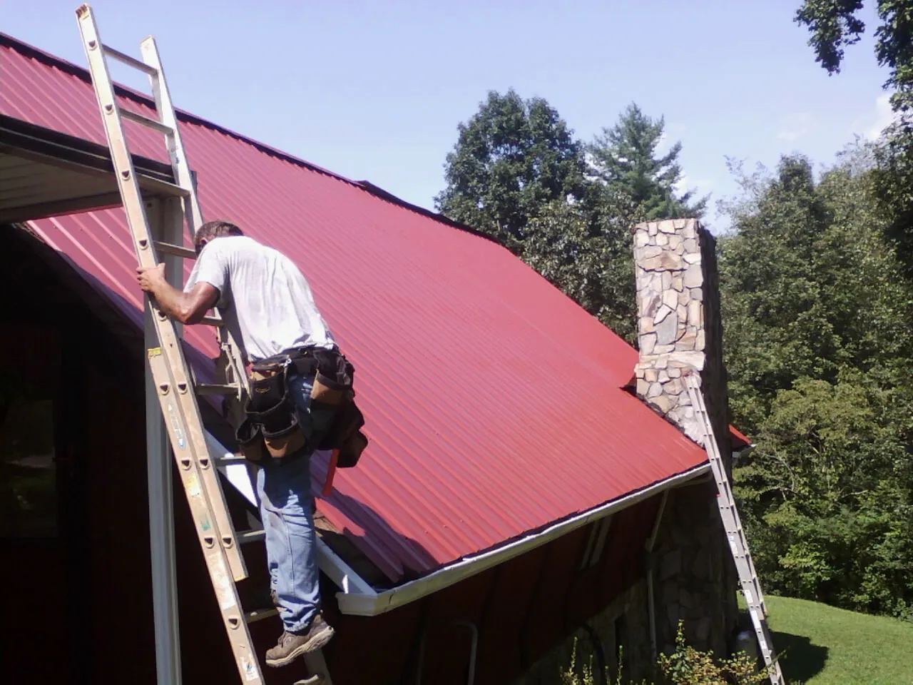 Roofer on a ladder beside a red metal roof and stone chimney — Soffit & Fascia Repair by Roofing Brothers in Memphis